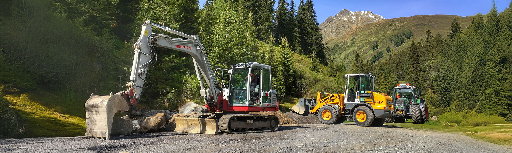 Bagger hebt Schotter auf Baustelle nahe Waldrand, mehrere Baumaschinen im Hintergrund, Berge bei klarem Himmel sichtbar.