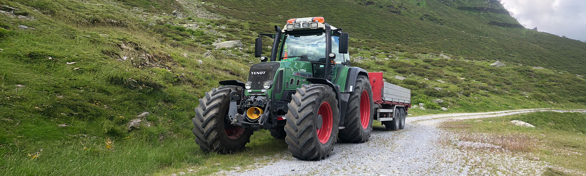 Traktor mit Anhänger fährt auf einem Schotterweg durch eine grüne Berglandschaft mit steinigem Untergrund.
