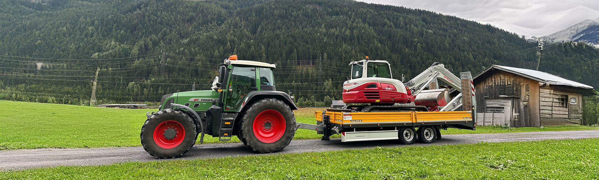 Traktor zieht Anhänger mit Bagger über eine schmale Straße durch grüne Wiesen, im Hintergrund Berge und eine Holzhütte.